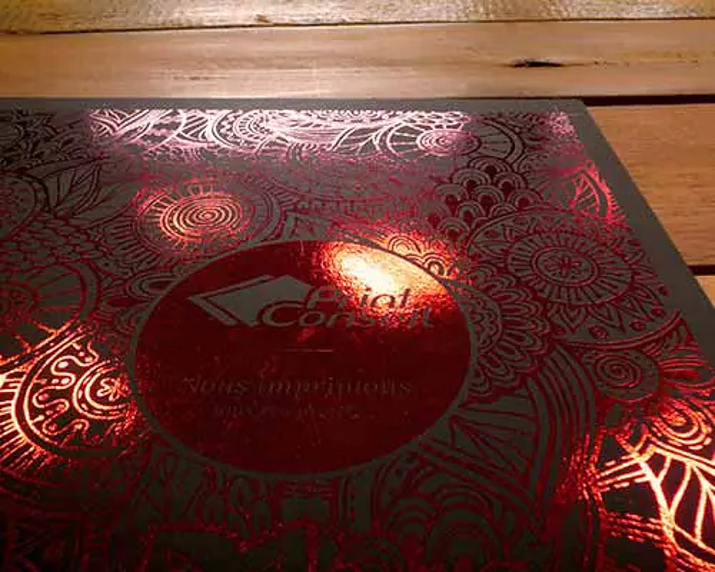 Une couverture noire ornée de motifs floraux rouge métallisé brillant et d'un logo central, posée sur une table en bois.