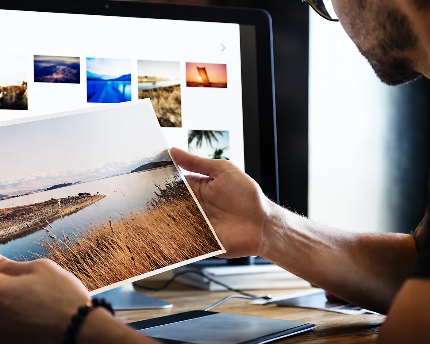 Un homme à lunettes tient un tirage photo d’un paysage de roseaux au bord de l’eau devant un écran affichant des miniatures de photos.