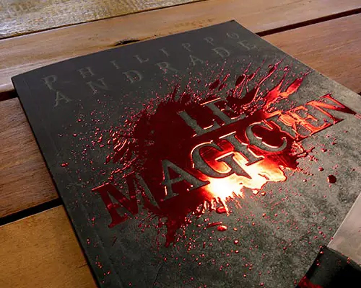 Couverture noire du livre Le Magicien de Philippe Andrade, ornée d'une éclaboussure rouge brillante, posée sur une table en bois.
