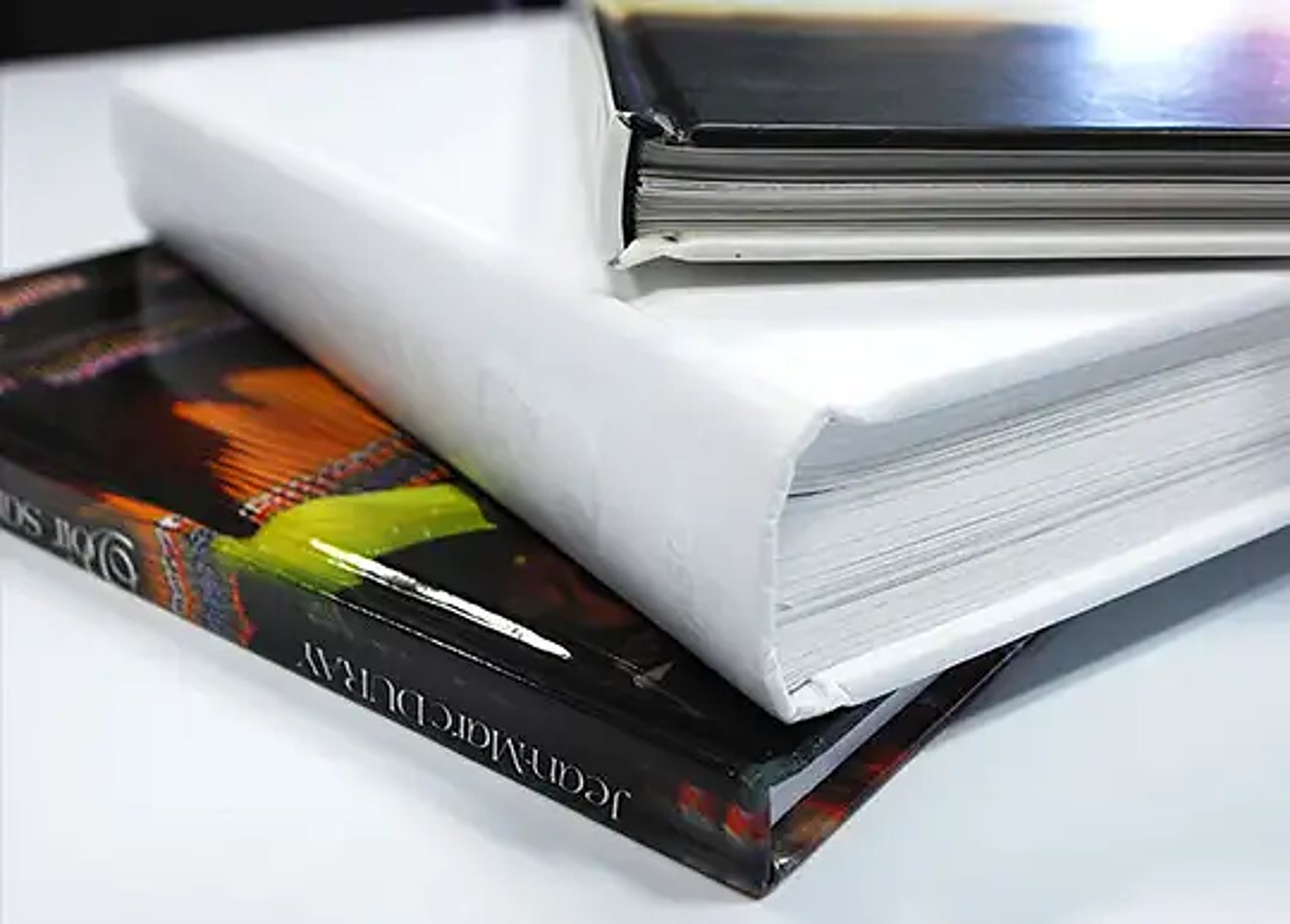 Trois livres à couverture rigide empilés sur une surface blanche, dont un livre blanc épais au centre et un livre illustré coloré en dessous.